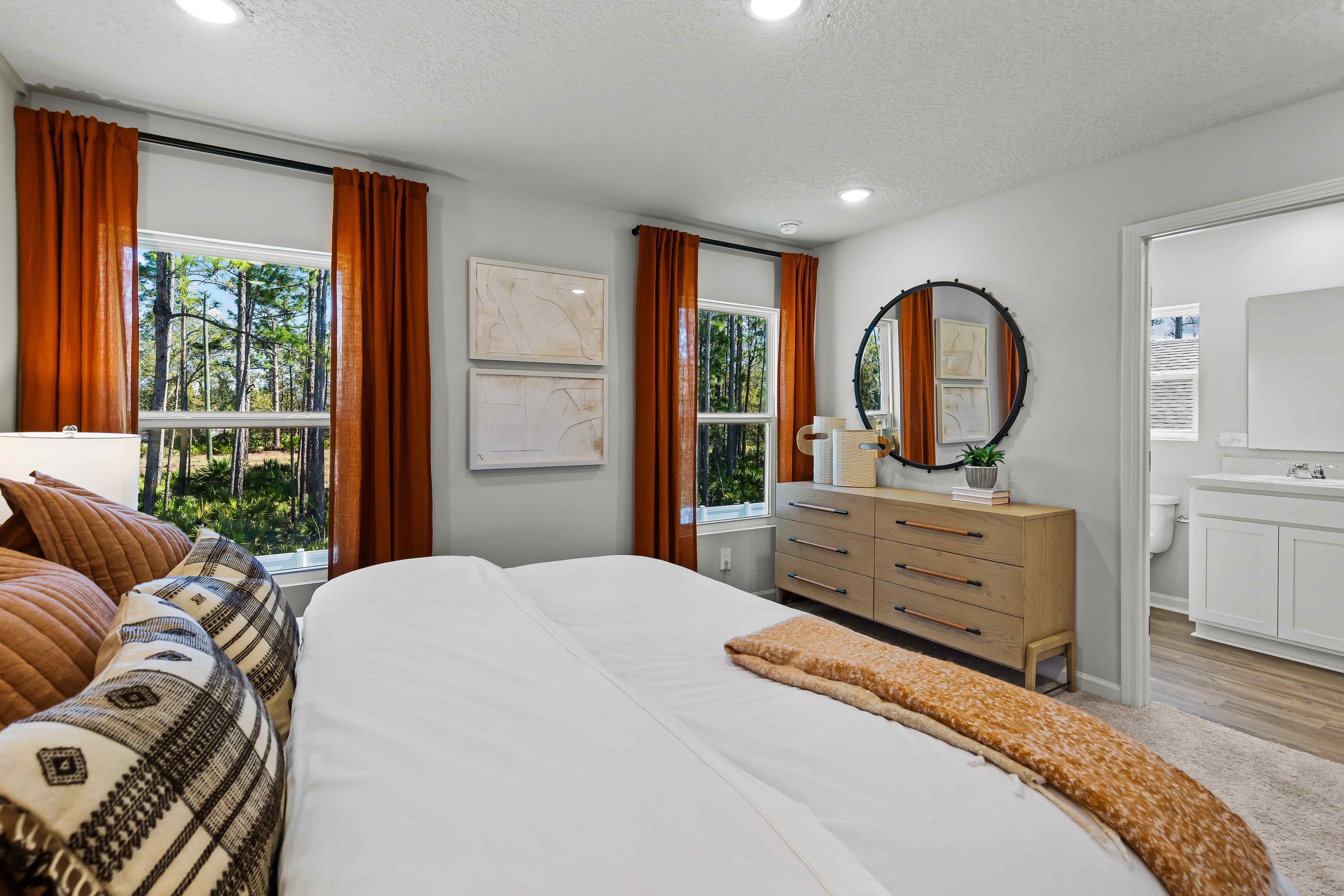 Davor Model Paired Home Primary Bedroom with Orange Bed Comforter and Two Windows with Curtains at The Landings at Pecan Park